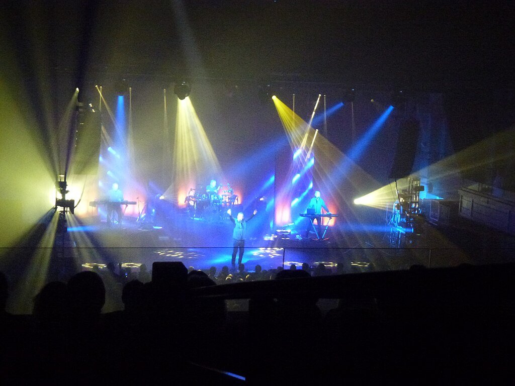 OMD se apresentando ao vivo no palco do Cambridge Corn Exchange em 2017, com iluminação azul e teclados em destaque